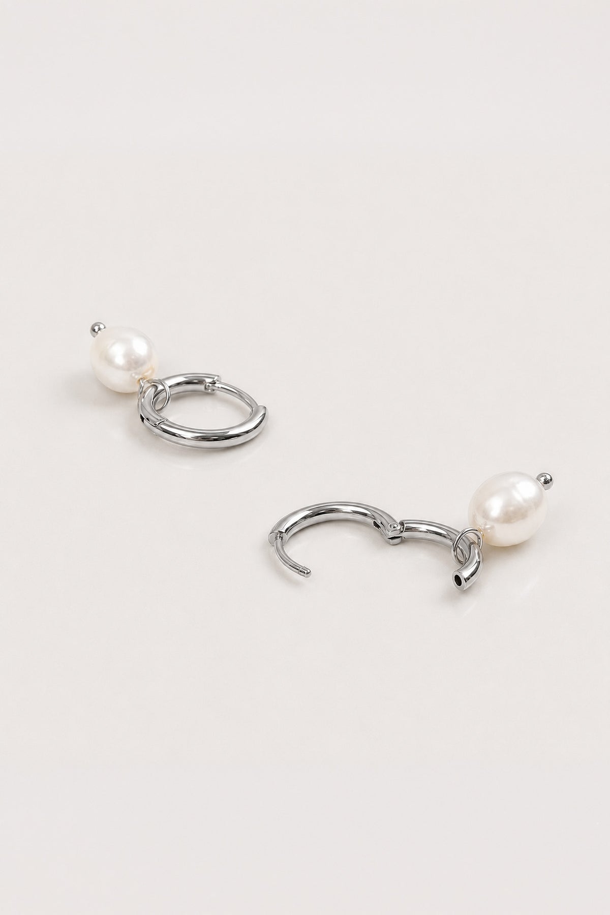 A pair of silver-toned hoop earrings with a small white pearl dangling below each hoop, displayed on a clean, light off-white background.