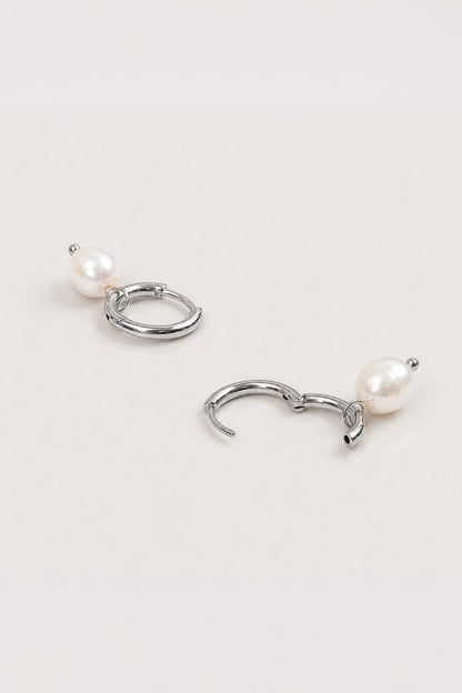 A pair of silver-toned hoop earrings with a small white pearl dangling below each hoop, displayed on a clean, light off-white background.