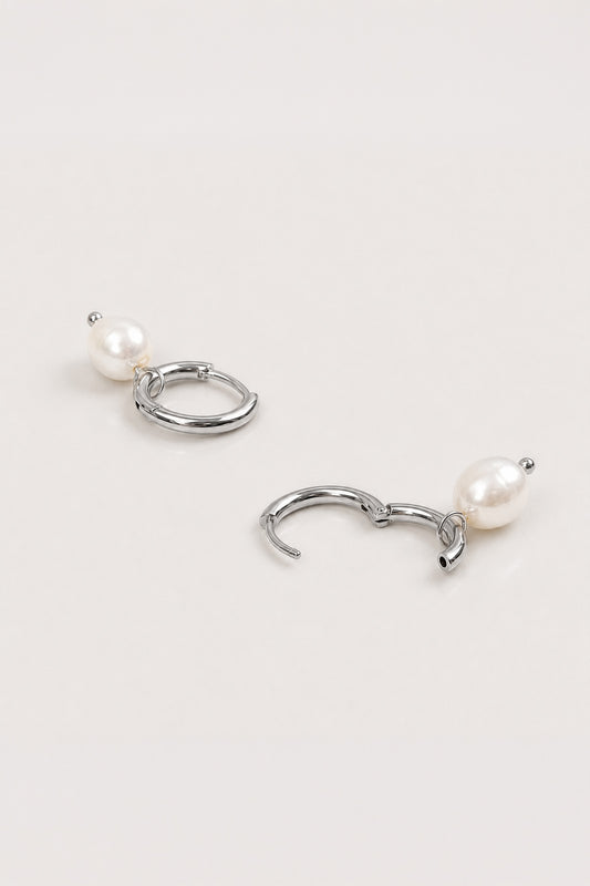 A pair of silver-toned hoop earrings with a small white pearl dangling below each hoop, displayed on a clean, light off-white background.